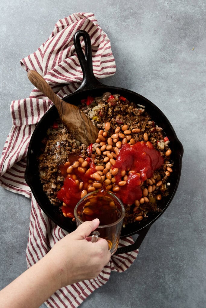 Delicious ground beef chili cooking in a cast iron skillet topped with beans and tomato sauce, with a wooden spoon and a hand pouring broth, on a red-striped cloth background.