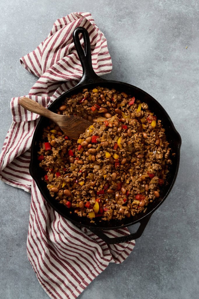 Delicious homemade beef chili with beans, colorful peppers, and spices cooked in a cast iron skillet, served on a gray surface with a red and white striped towel, perfect for comforting meals and family dinners.