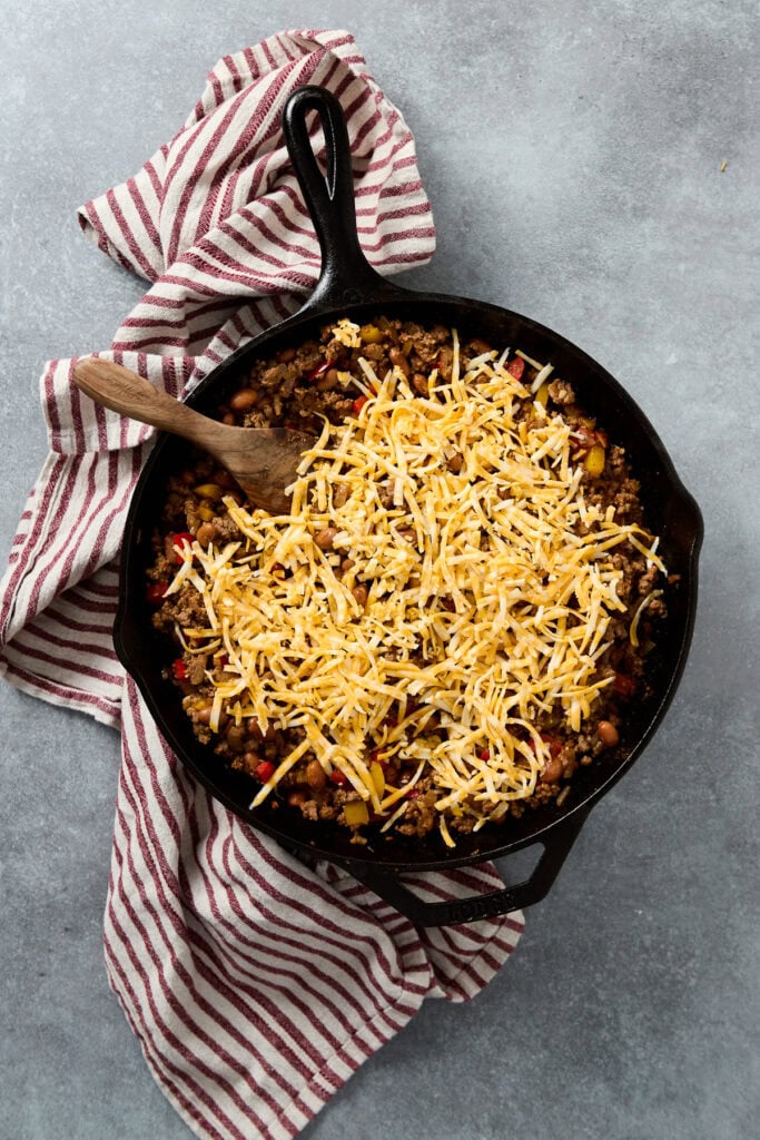 Cheddar cheese melting over seasoned ground beef and pasta in a cast iron skillet, topped with shredded cheese, served on a rustic cloth for a comforting meal.
