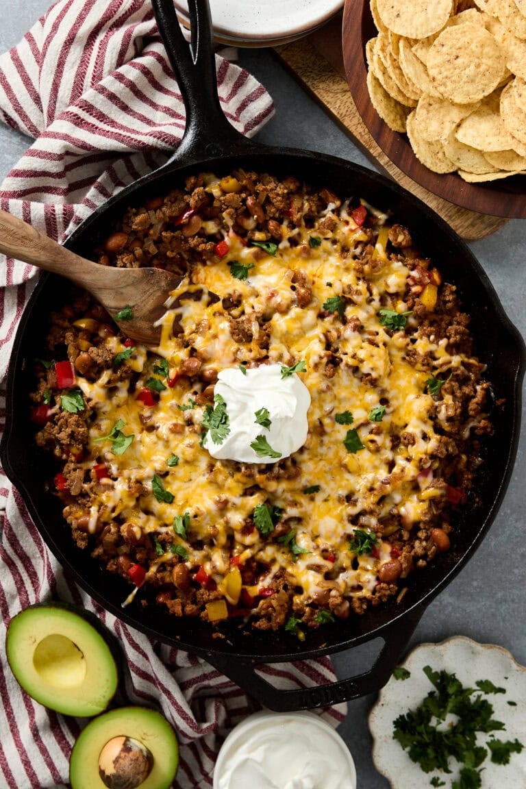 A delicious easy beef taco skillet topped with melted cheese, seasoned ground beef, fresh cilantro, and a dollop of sour cream, served with avocado slices and crispy tortilla chips on the side. Perfect for snack or party food.