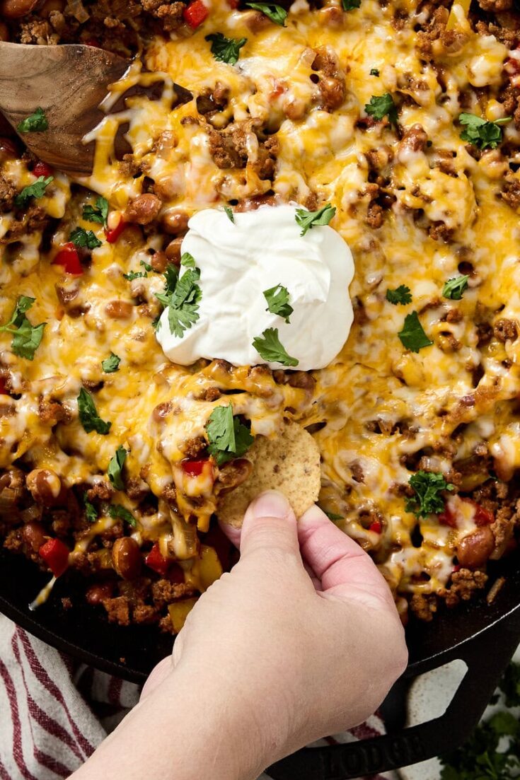 Delicious beef taco skillet with melted cheese, ground beef, beans, and vibrant red peppers, garnished with fresh cilantro and a dollop of sour cream, served with crunchy tortilla chips.