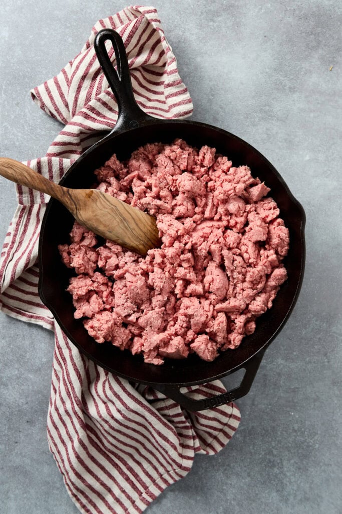 Alt text: Cooked ground beef in a cast-iron skillet with a wooden spoon, on a gray surface with a striped kitchen towel, perfect for meat recipes.