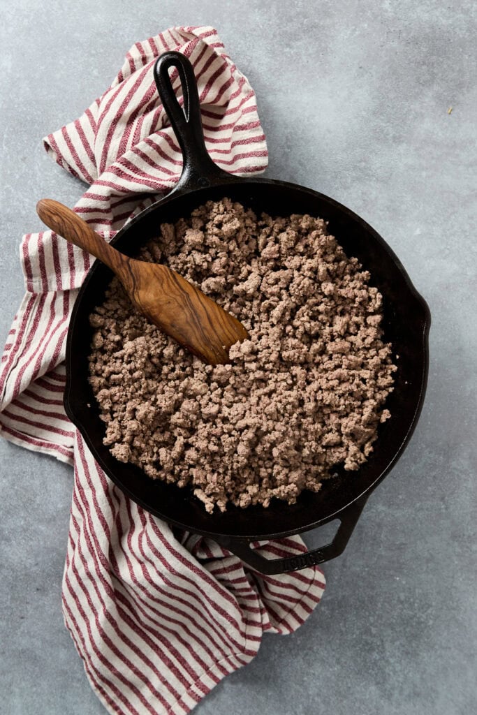 Alt text: Cast iron skillet with cooked ground beef, wooden spatula, red and white striped cloth, on gray surface, perfect for recipes and cooking tips.