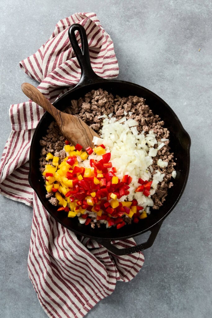 A cast iron skillet filled with cooked ground beef, chopped onions, and diced red, yellow bell peppers, ready for a tasty meal.