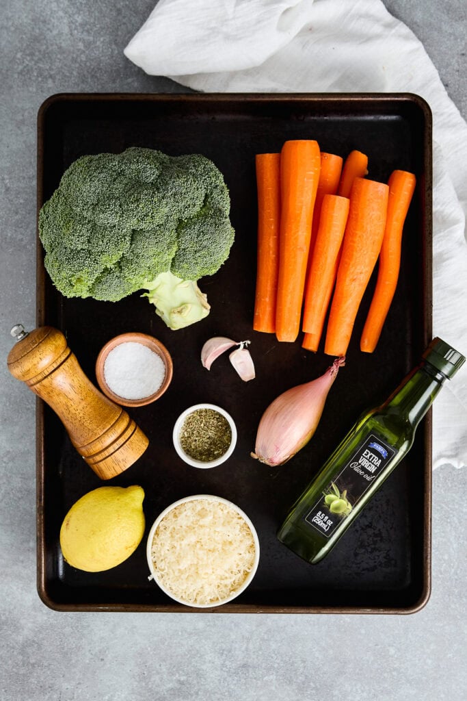 Brightly colored fresh broccoli, carrots, garlic, lemon, and seasonings on a baking tray.