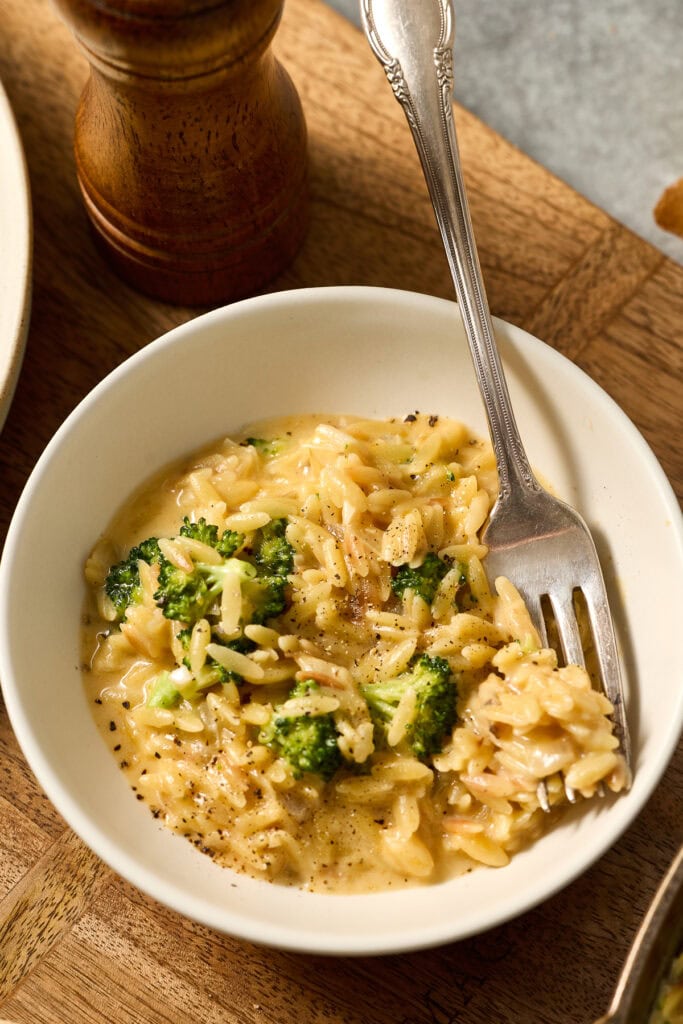Delicious creamy broccoli cheddar orzo served in a white bowl with a fork, garnished with black pepper, on a wooden table. Perfect comfort food for all ages.