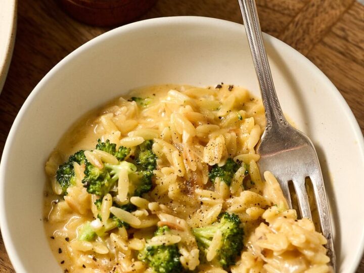 Delicious creamy broccoli cheddar orzo served in a white bowl with a fork, garnished with black pepper, on a wooden table. Perfect comfort food for all ages.