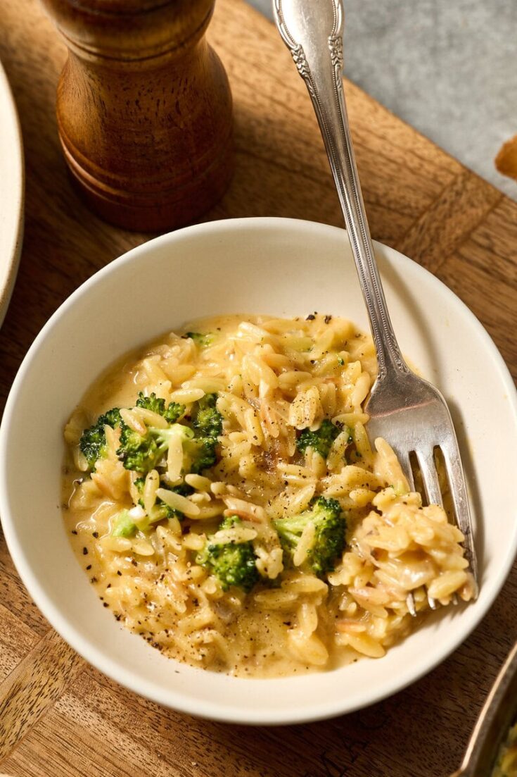 Delicious creamy broccoli cheddar orzo served in a white bowl with a fork, garnished with black pepper, on a wooden table. Perfect comfort food for all ages.