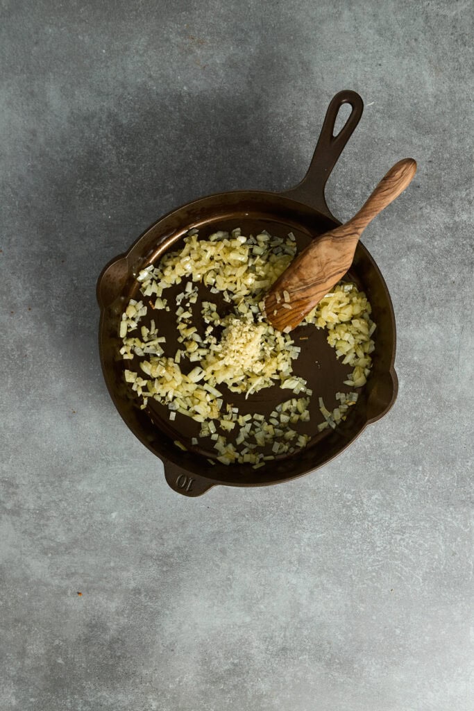 Sautéed garlic and onions in a cast iron skillet with a wooden spoon, ready for cooking or serving, on a gray textured surface.