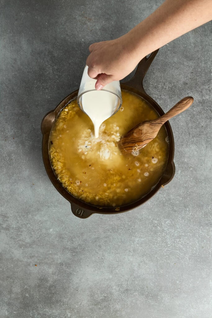 Creamy broccoli cheddar orzo being prepared in a skillet with milk being poured in.