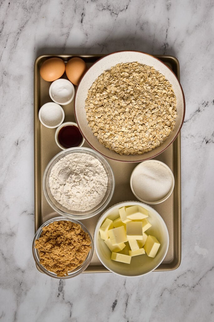 Ingredients for soft and chewy brown butter oatmeal cookies including oats, butter, eggs, flour, sugar, and brown sugar on a baking tray.
