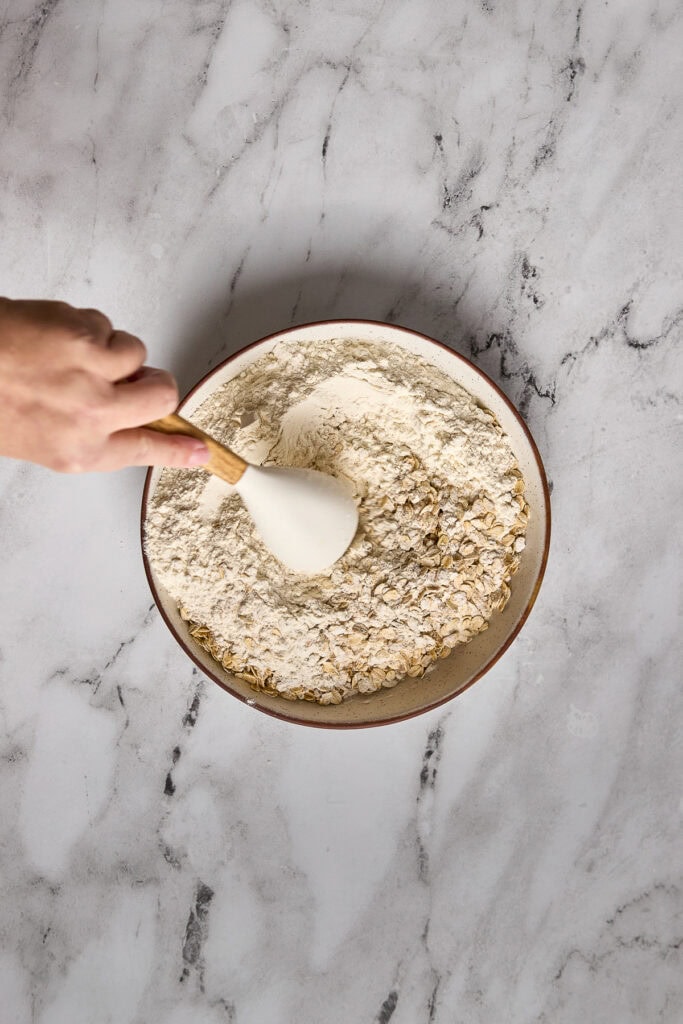 Bowl of dry oatmeal cookie ingredients with a hand holding a spoon, ready for mixing, on a marble surface. Perfect for baking soft and chewy brown butter oatmeal cookies.