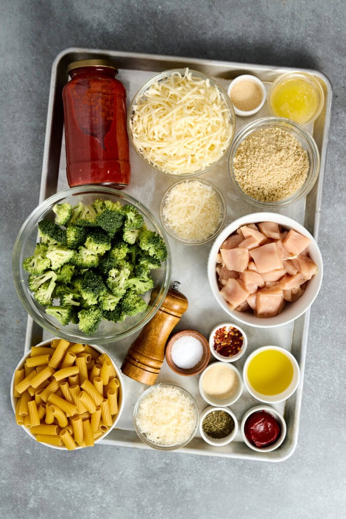 Assorted ingredients for Chicken Parmesan skillet including broccoli, shredded cheese, pasta, chicken, and seasonings on a metal tray.