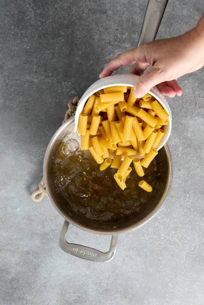 Hand pouring uncooked rigatoni pasta into a skillet with boiling water, ready for Chicken Parmesan recipe preparation.