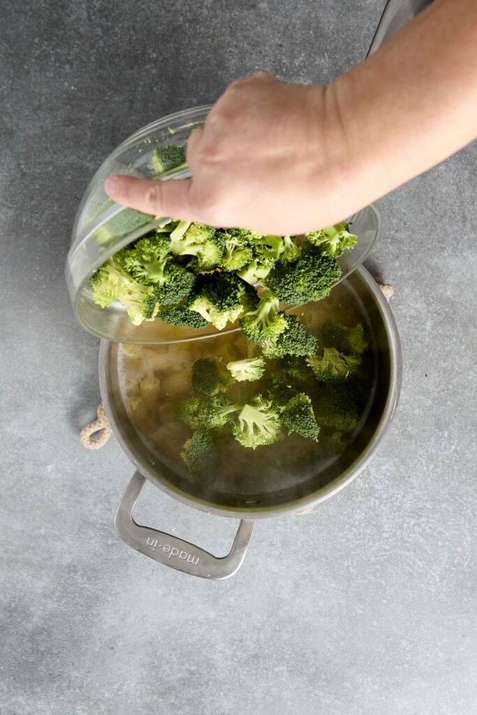 Hand pouring fresh broccoli florets into a pot of boiling water, preparing vegetables for Chicken Parmesan Skillet recipe.