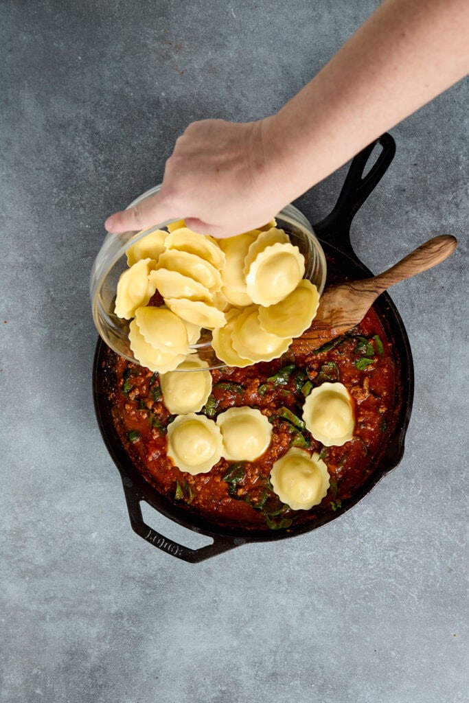 Fresh ravioli being placed into a skillet with hearty tomato sauce, highlighting homemade pasta and Italian cooking.