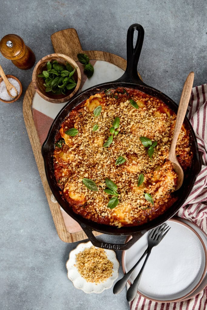 Delicious ground beef and spinach ravioli in a cast iron skillet topped with melted cheese and fresh basil, served with side dishes and utensils on a rustic table.