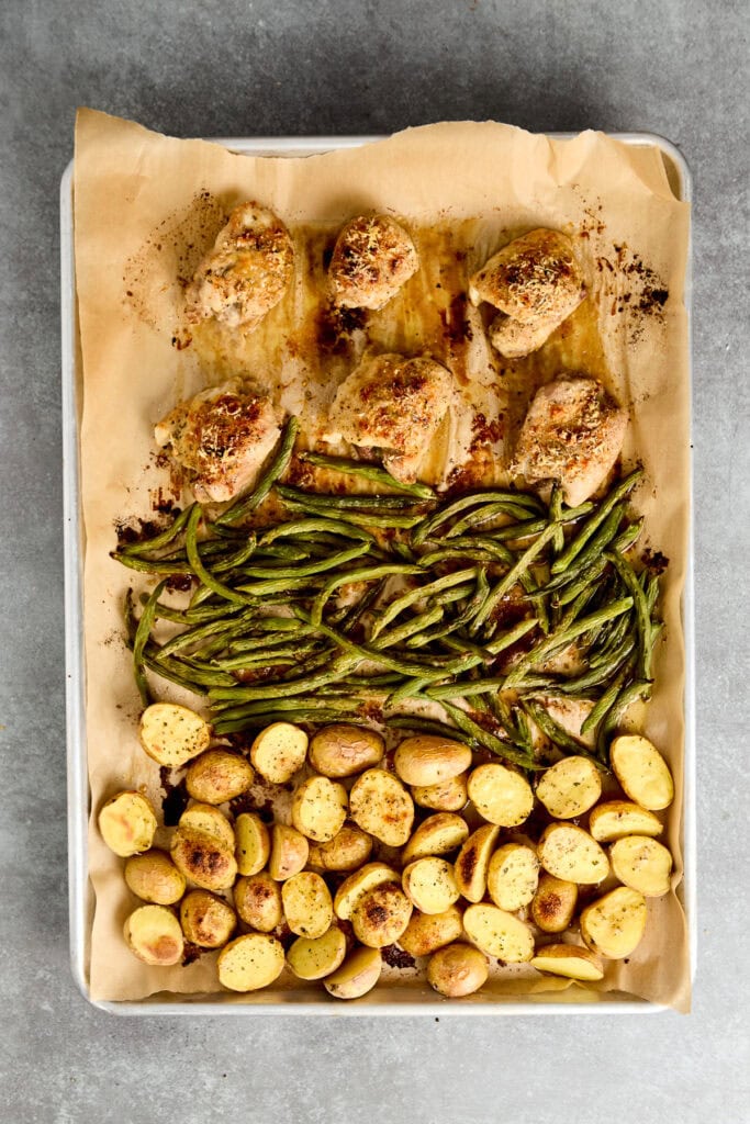 Baked chicken thighs with roasted green beans and golden potatoes on parchment paper, served on a baking tray.