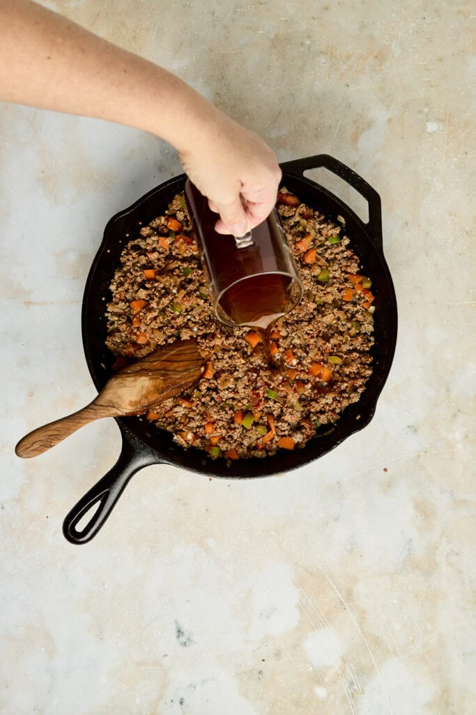 Cooking ground beef with vegetables in a cast iron skillet, adding spices for flavor, on a light-colored surface.