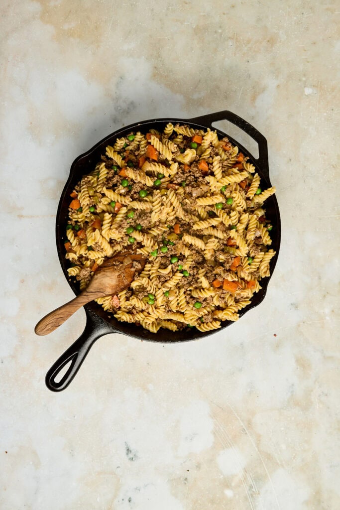 Colorful vegetable pasta salad in a black skillet with a wooden spoon, featuring rotini pasta, carrots, peas, and herbs, perfect for a fresh, healthy meal.
