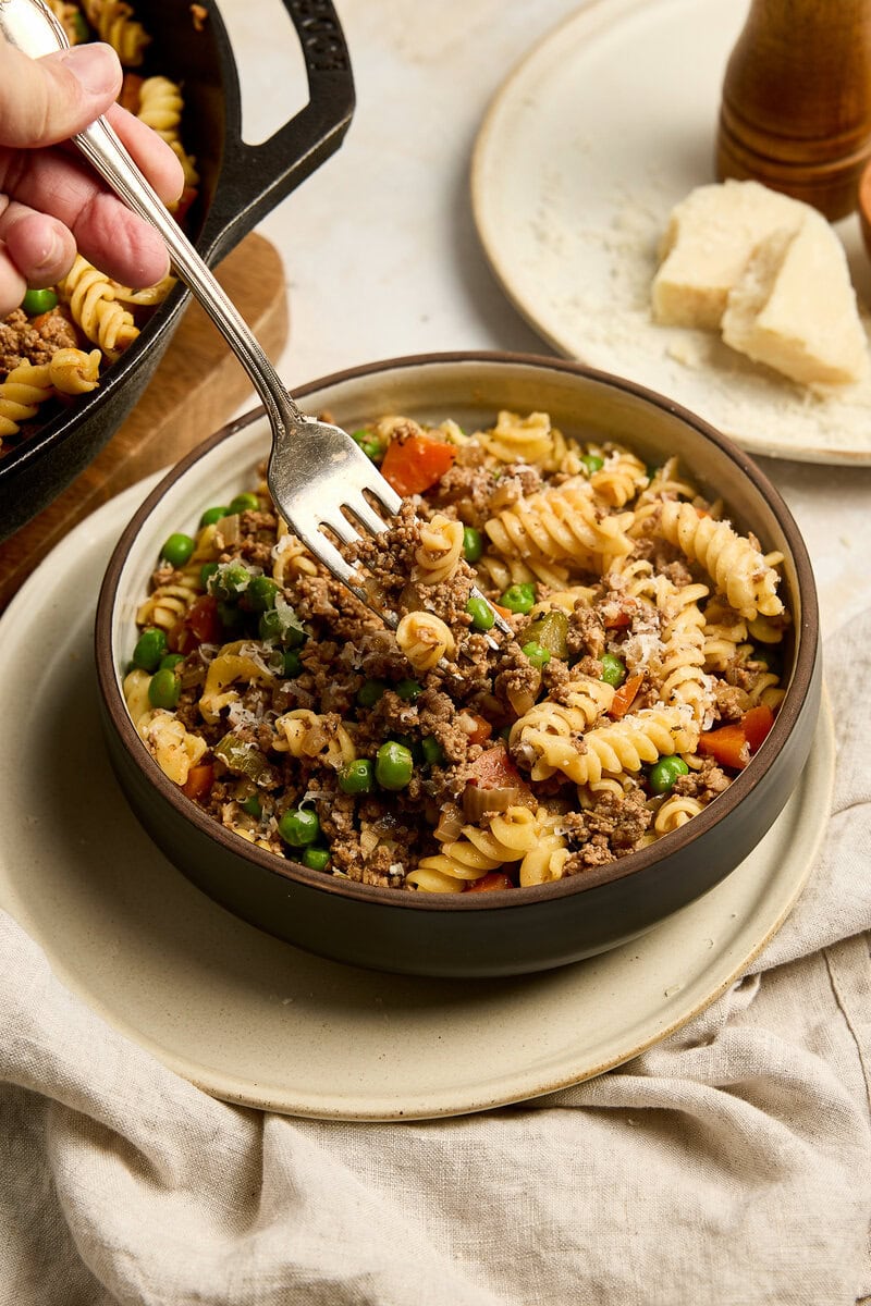 Hearty ground beef pasta with vegetables served in a black bowl, garnished with herbs, on a neutral table setting. Perfect comfort food for family dinners.