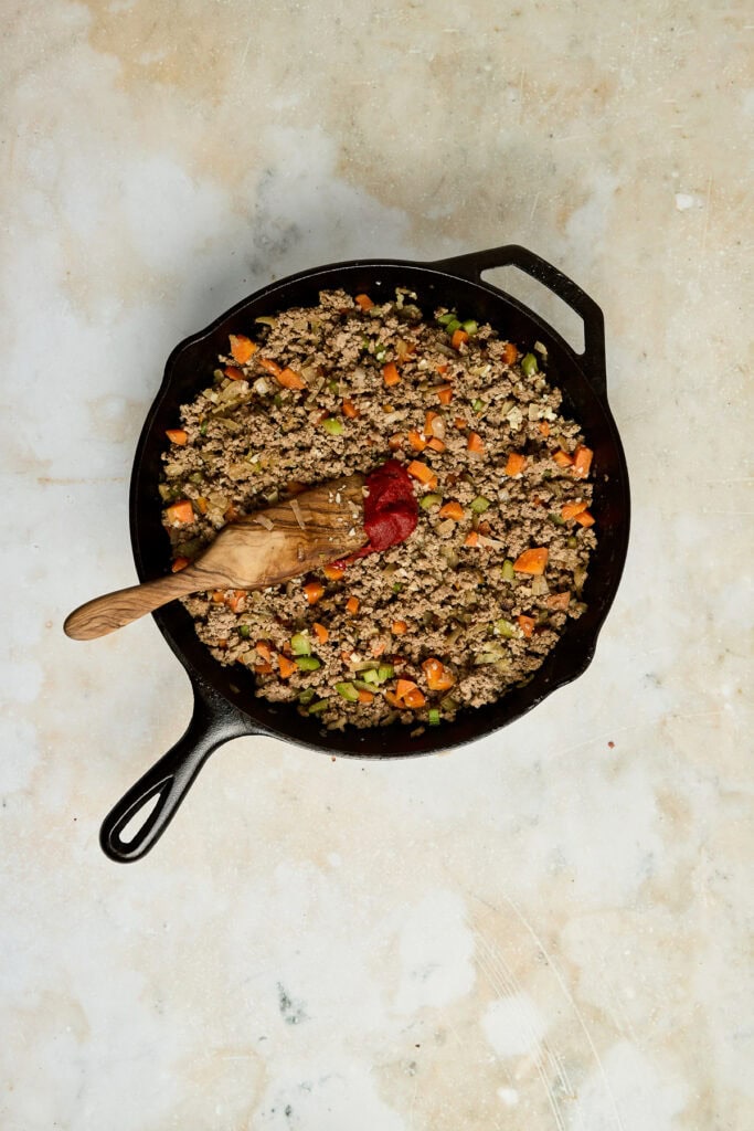 Ground beef and vegetable skillet in a cast iron pan with a wooden spoon, featuring cooked ground beef, carrots, and green peas, on a rustic surface.