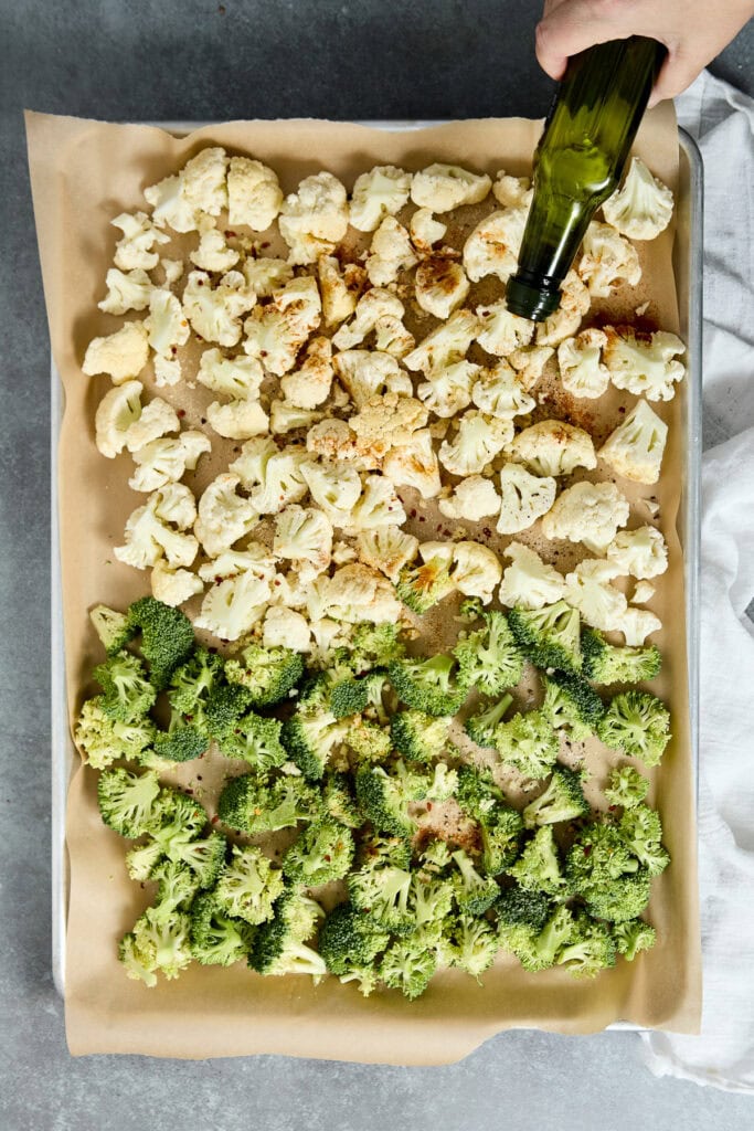 Roasted cauliflower and broccoli florets on a baking sheet, with a hand pouring olive oil over the vegetables, perfect for healthy snacking and meal prep.