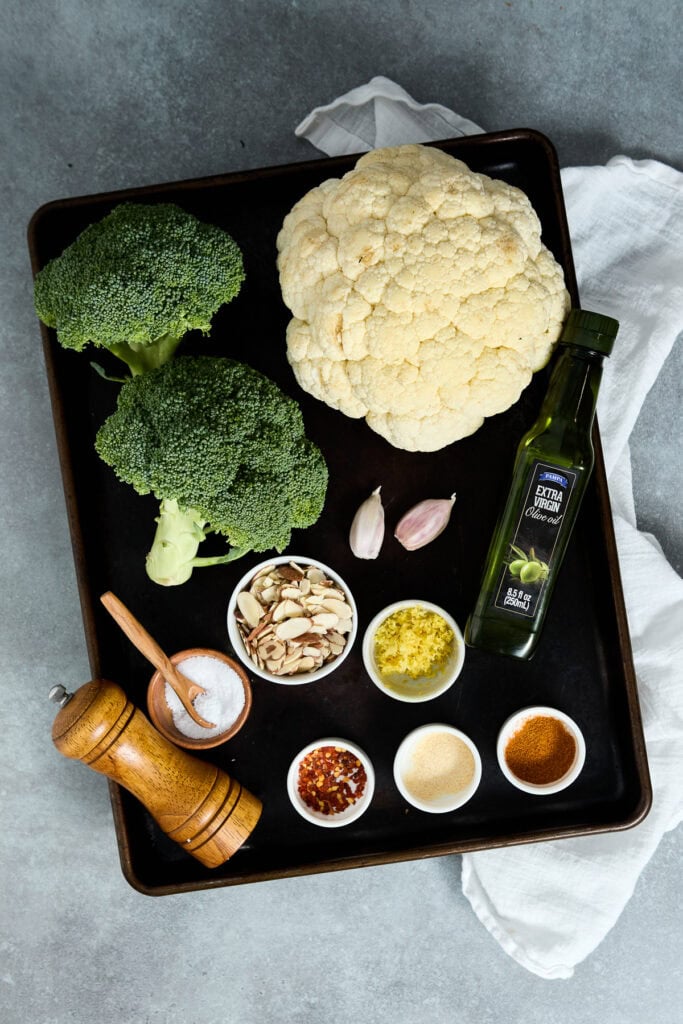 Fresh cauliflower and broccoli with garlic, spices, and oil on a black tray, ready for cooking or recipe preparation.