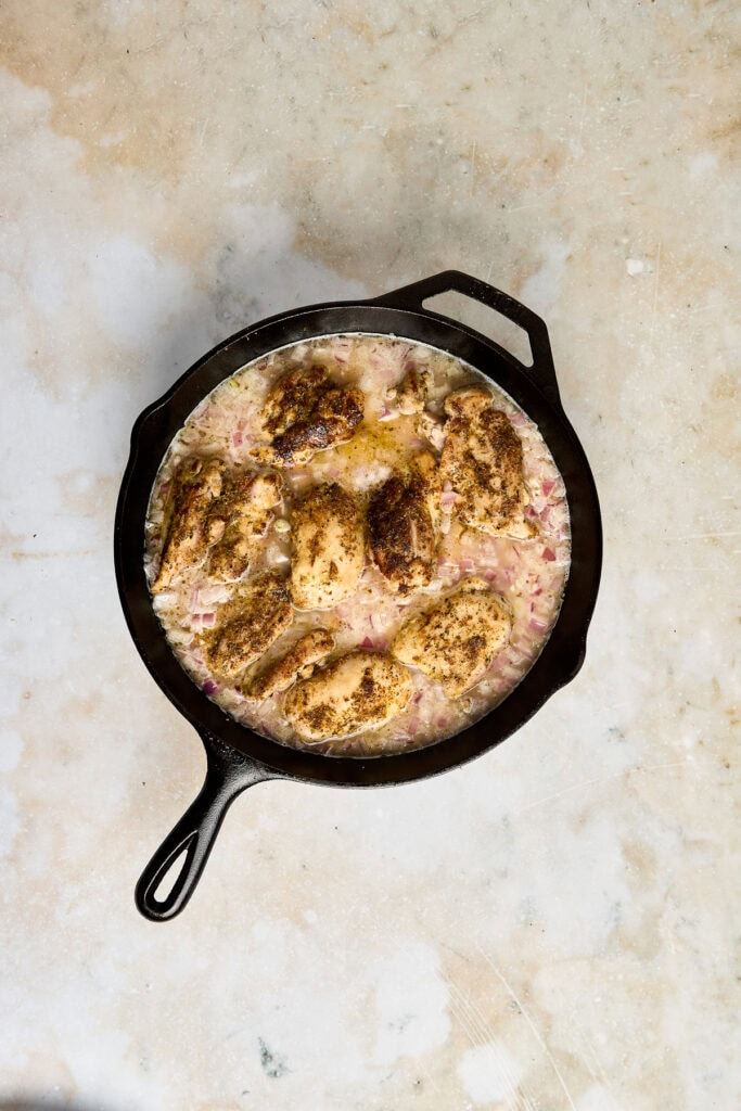 Delicious greek chicken thighs cooking with lemon rice in a black cast iron skillet, featuring golden-brown crust and tender, moist interior. Perfect comfort food for breakfast, brunch, or dinner.