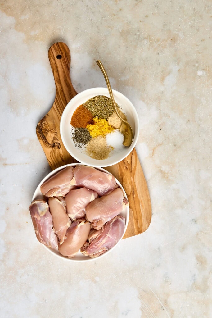 Raw chicken thighs arranged on a white plate with a bowl of mixed spices and herbs, ready for seasoning and cooking, on a rustic wooden cutting board.