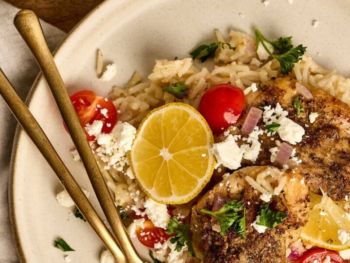 Juicy greek chicken with lemon slices, cherry tomatoes, and rice garnished with herbs on a white plate. Perfect for a flavorful, healthy meal.
