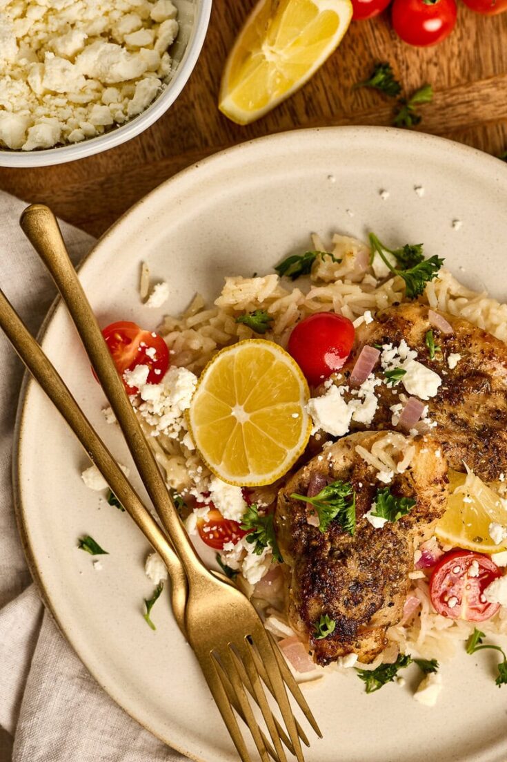 Juicy greek chicken with lemon slices, cherry tomatoes, and rice garnished with herbs on a white plate. Perfect for a flavorful, healthy meal.