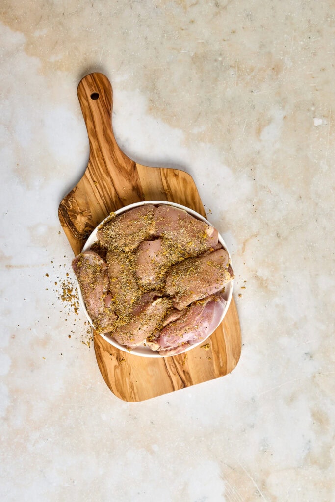 Raw chicken cutlets coated with seasoned breading, ready for cooking, placed on a round white plate on a rustic wooden cutting board.