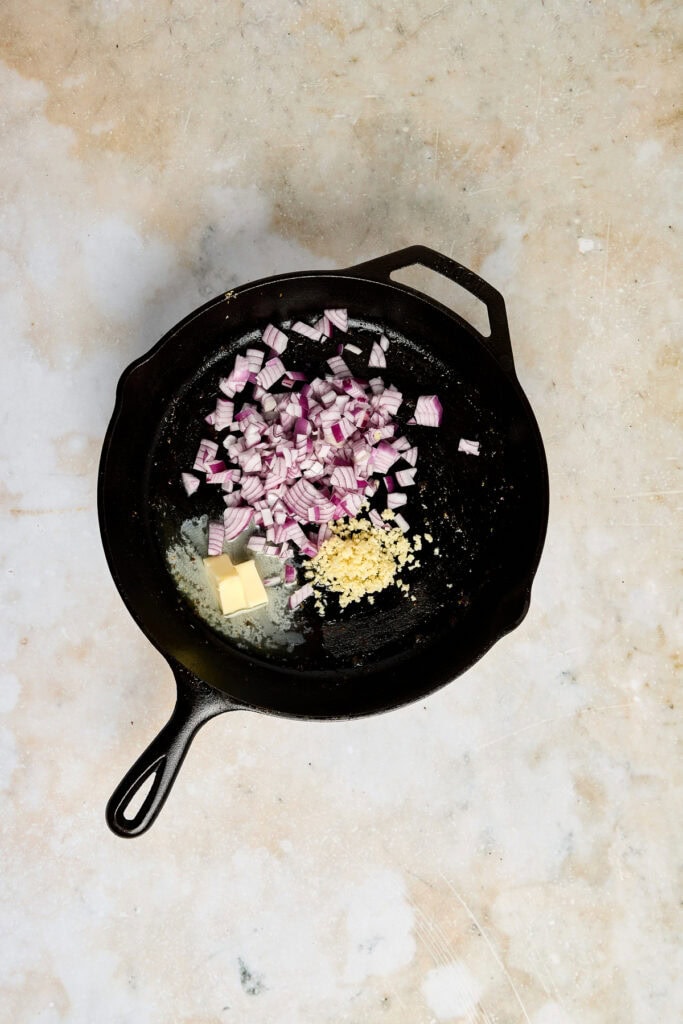 Cast iron skillet with chopped red onions, minced garlic, and butter ready for cooking, on a textured beige surface.