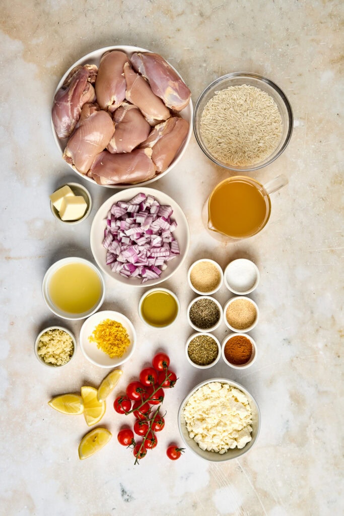 Raw chicken thighs, rice, and fresh vegetables arranged on a light surface, ready for cooking. Includes garlic, lemon, cherry tomatoes, and spices for flavorful meal preparation.