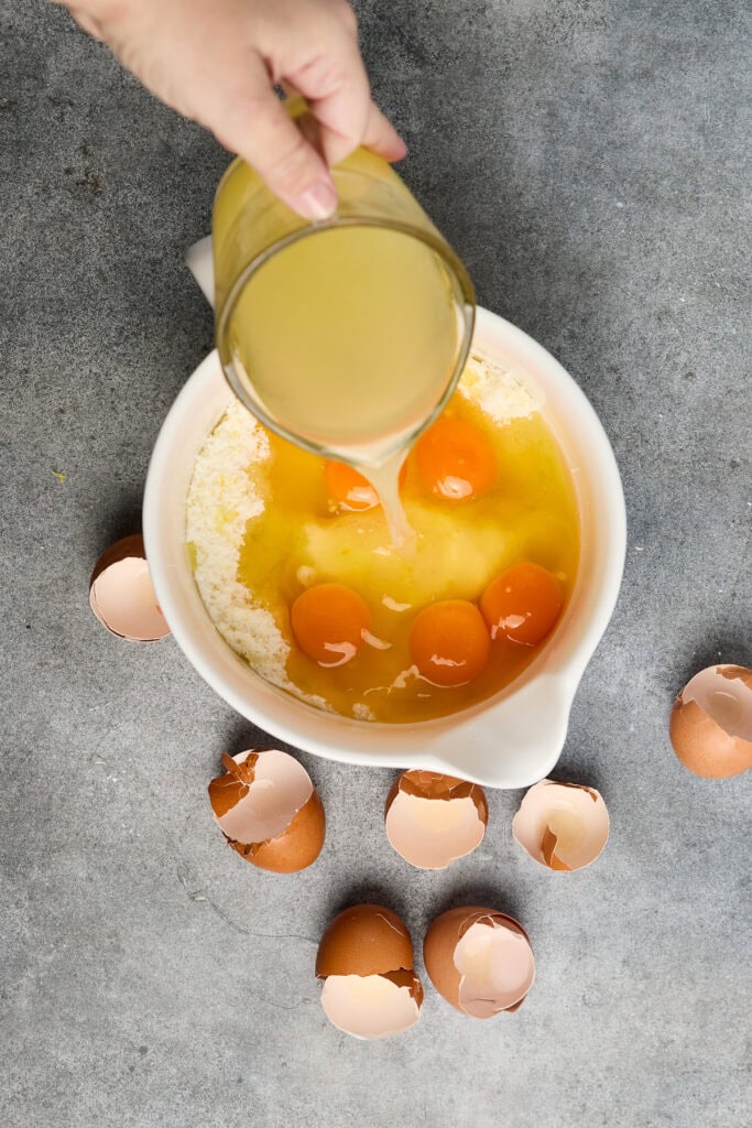 Pouring liquid ingredients into a bowl of cake batter with eggs, ready for baking.