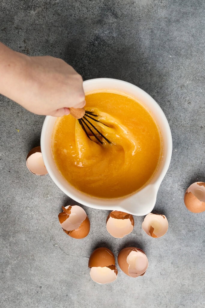 Hand whisking a vibrant yellow egg mixture in a white bowl, surrounded by cracked eggshells on a gray surface, perfect for baking or dessert preparation.