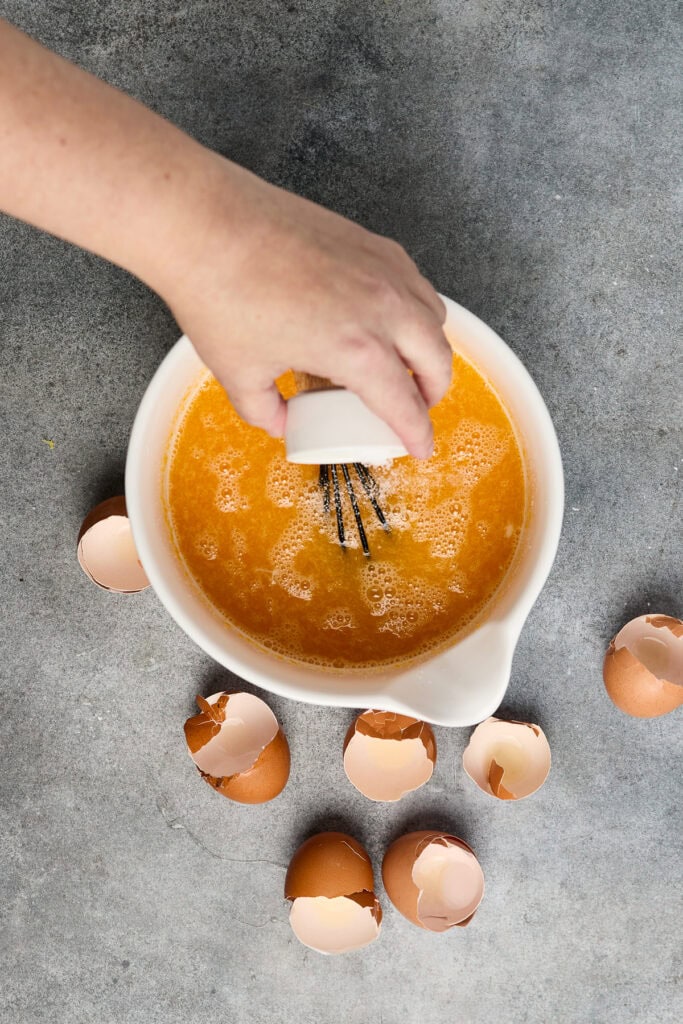 Hand whisking fresh orange juice in a white pitcher surrounded by eggshells on a gray surface, emphasizing fresh ingredients and homemade beverage preparation.