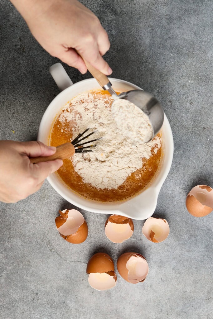 Close-up of hands mixing cake batter with flour and eggs in a white bowl, surrounded by cracked eggshells on a gray surface.