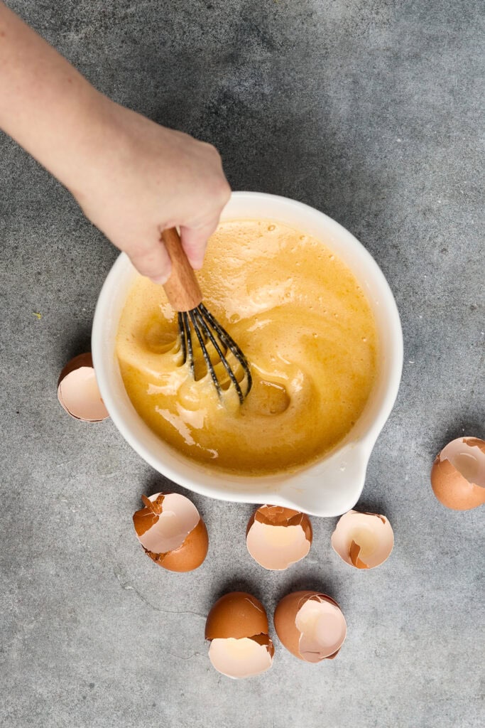 Hand whisking cake batter in a bowl with cracked eggshells around, ready for baking. Perfect for baking enthusiasts and dessert lovers.