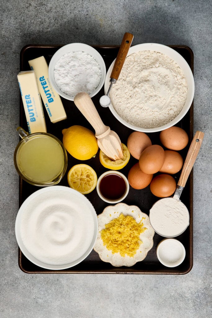Assorted baking ingredients including flour, eggs, butter, lemon, and sugar arranged on a baking tray for lemon bar preparation.