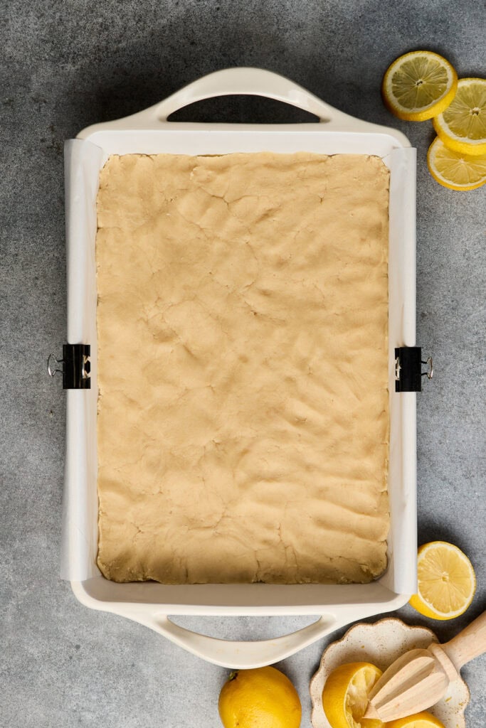 Freshly prepared lemon bar dough in a baking pan, ready for baking, with lemon slices and a citrus juicer nearby, perfect for making sweet and tangy lemon desserts.