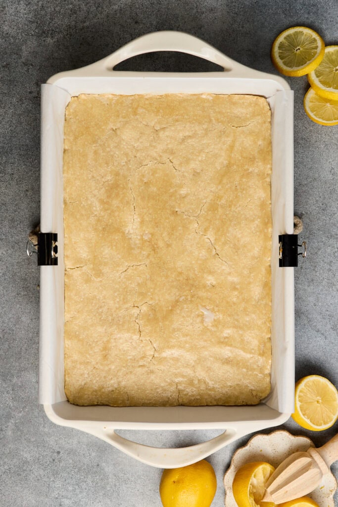 Freshly baked lemon bars on a white baking tray with lemon slices and a lemon juicer, perfect for citrus dessert lovers.