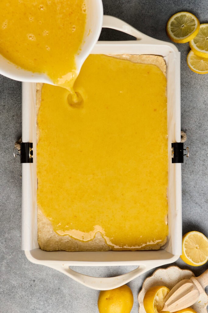 Freshly poured lemon filling over a baked lemon bar in a white baking dish, with lemon slices and a juicer nearby, perfect for a sweet and tangy dessert.