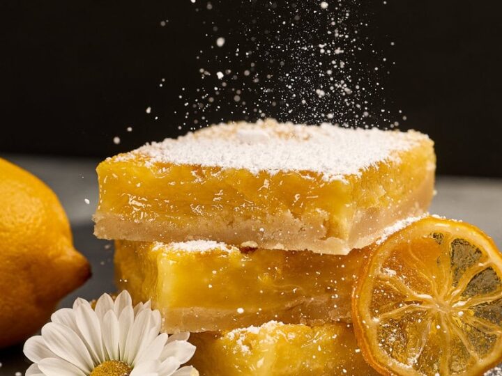 Lemon bars dusted with powdered sugar, fresh lemon slices, and a daisy flower, showcasing a sweet and tangy dessert perfect for lemon lovers.