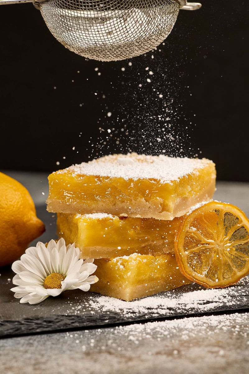 Lemon bars dusted with powdered sugar, fresh lemon slices, and a daisy flower, showcasing a sweet and tangy dessert perfect for lemon lovers.