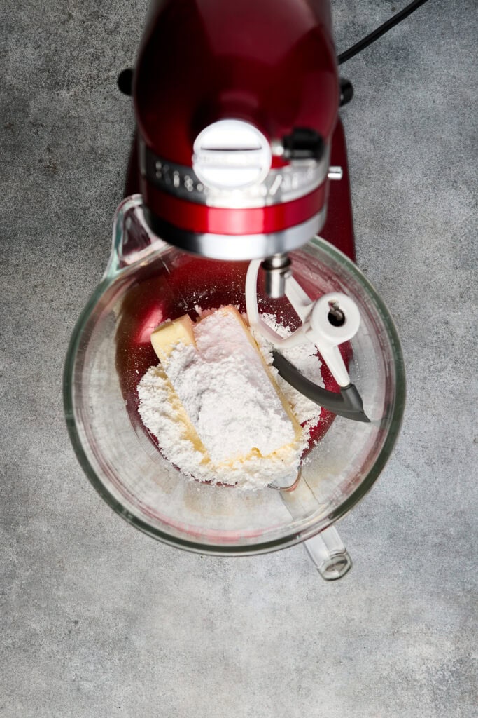 View of a red stand mixer with a glass bowl containing cake batter ingredients, including flour and butter, ready for baking.