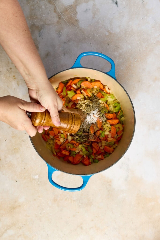 Person seasoning a colorful vegetable and rice mixture in a blue pot, preparing Lemon Chicken Rice Soup with fresh ingredients.