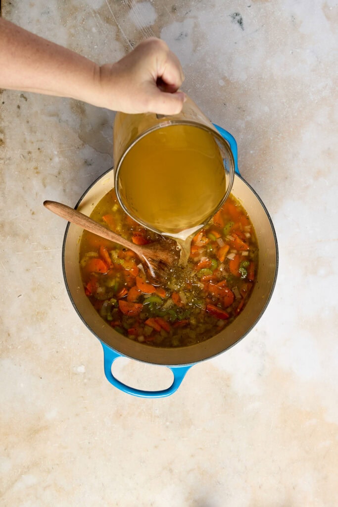 Pouring broth into a pot of lemon chicken rice soup with vegetables, on a neutral countertop.