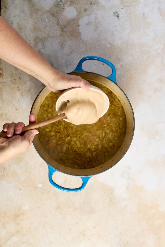 Person adding creamy sauce to a pot of lemon chicken rice soup, showcasing a step in the cooking process.
